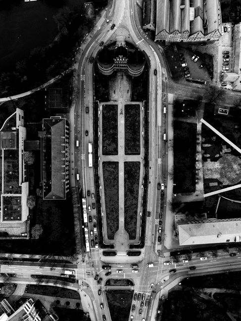 Aerial black-and-white photograph of a city street scene featuring a central landscaped park with symmetrical flower beds and paved walkways, surrounded by multi-story buildings and roads filled with 