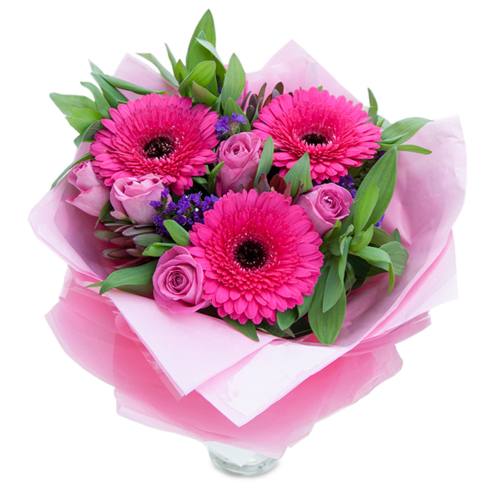 I often think of pink bouquets as the floral equivalent of a warm smile, and this one is no exception. Three fuchsia Gerberas dominate the scene with vibrancy-each daisy's petals arranged in a perfect radial pattern around a dark centre that grounds the palette. Between them, dusty lavender-pink roses open slowly, their layered petals creating soft, romantic volumes that invite touch. Scattered like tiny wild accents are clusters of purple statice, their papery texture and cool hue introducing contrast and depth. A generous spray of deep green foliage-broad, glossy leaves resembling salal with occasional eucalyptus tones-wraps the blooms and lends a fresh, pastoral base that reminds me of bouquets sold at the weekend market by the Cray Valley. The wrapping is part of the composition: an inner sheet of pale pink tissue provides a matte, gentle bed, while an outer sheet of translucent fuchsia-pink cellophane, slightly crinkled, adds a luminous sheen and tactile interest. The top rim of a clear glass vase peeks out below, indicating the arrangement is arranged for immediate display on a coffee table or as a centrepiece. In soft daylight the colours read bright and optimistic, the fragrance subtle-roses offering a classic perfume, Gerberas a faint green undertone-and the combined textures create a balanced, uplifting presence. Crafted with local care, this bouquet is ideal for celebrating birthdays, marking anniversaries, or simply lifting someone's day across St Mary Cray and neighbouring Crayford and Orpington streets.